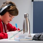 LearnOnline IGCSE Packages 14-16 year old boy with headphones on, writing in book in front of laptop