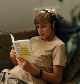 LearnOnline English IGCSE Package. Young teen sat on sofa reading a book with headphones on.